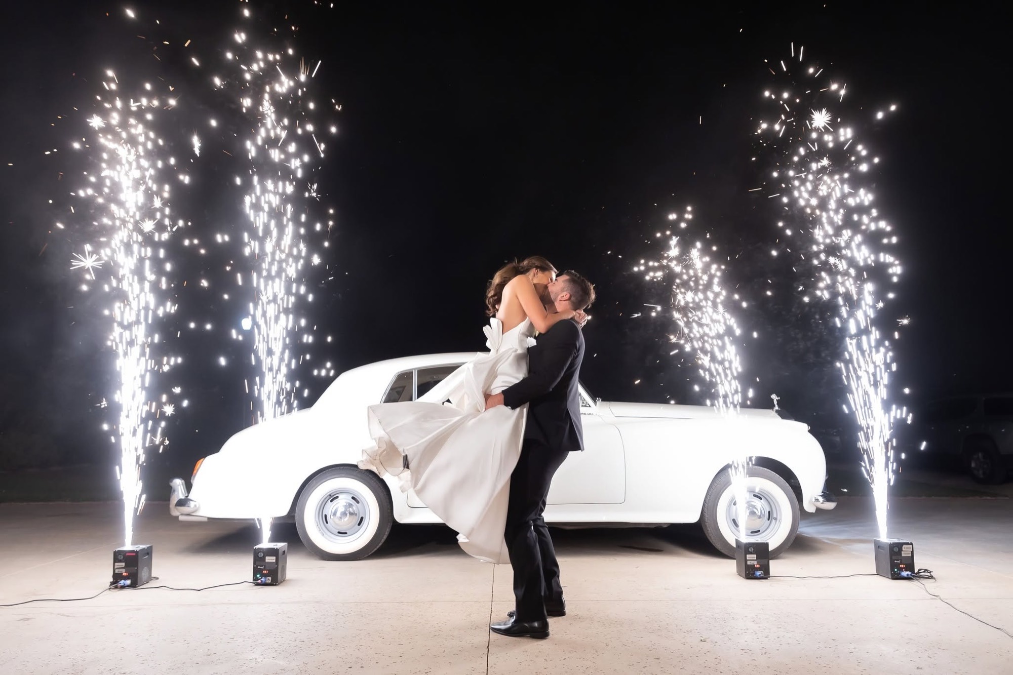 A bride and groom share a first kiss in front of a vintage white car with sparkler fountains at a CM Promotions wedding